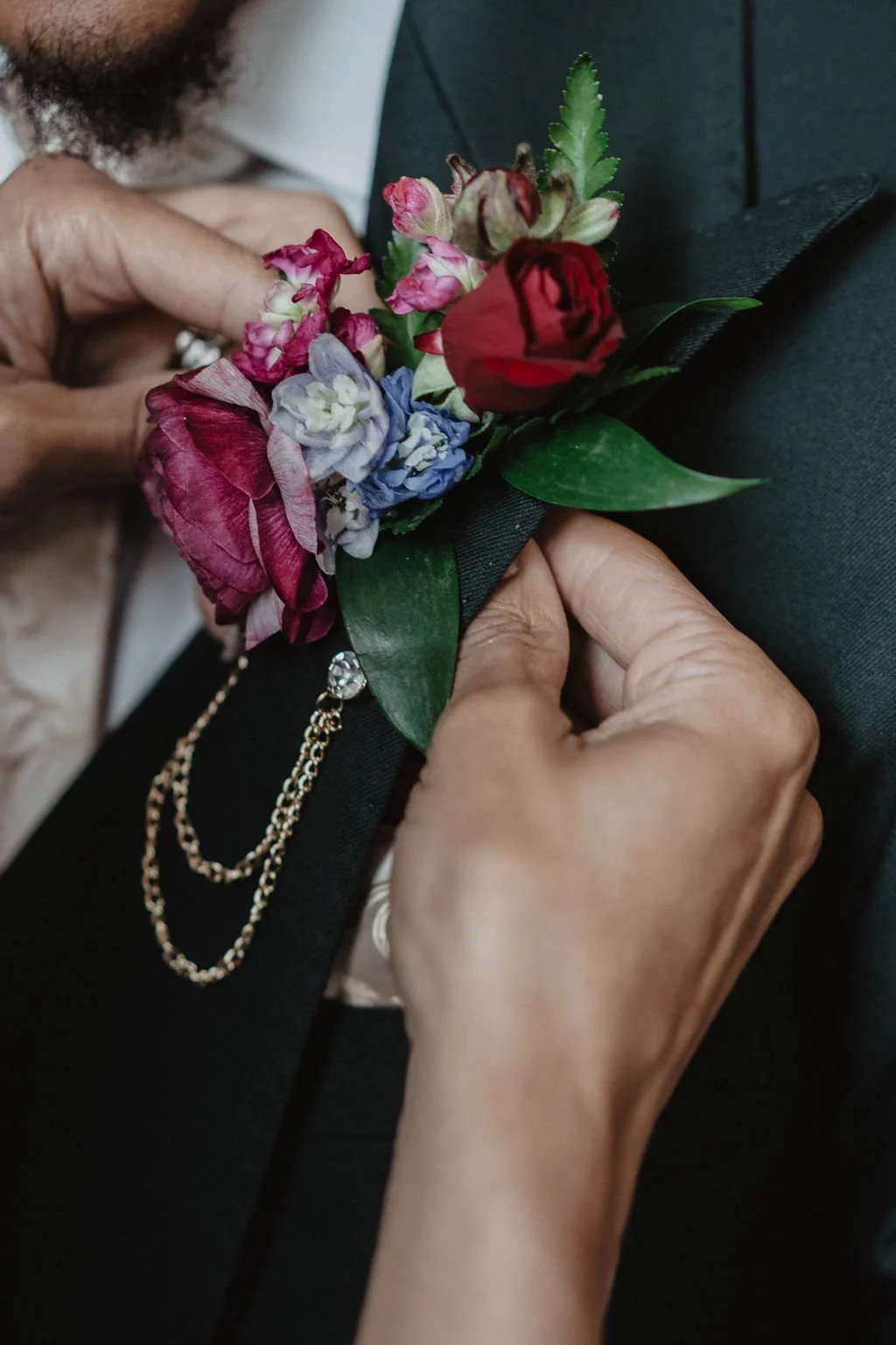 Close-up of a person pinning a colorful boutonniere with red, pink, purple, and white flowers, along with green leaves, onto black fabric.