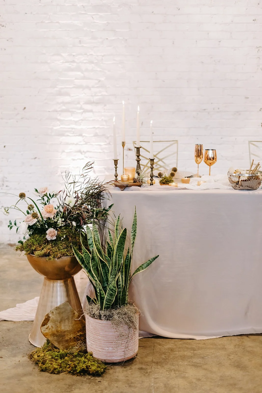 A decorated table with candles, rose gold glasses, and a variety of plants, including a pink flower arrangement and a green leafy plant, set against a white brick wall.