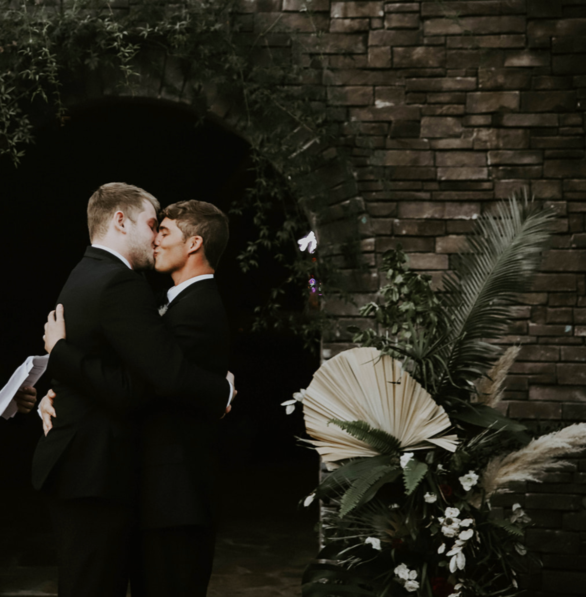 Two men in black suits sharing a kiss at a wedding ceremony, with a brick arch and floral arrangement in the background.
