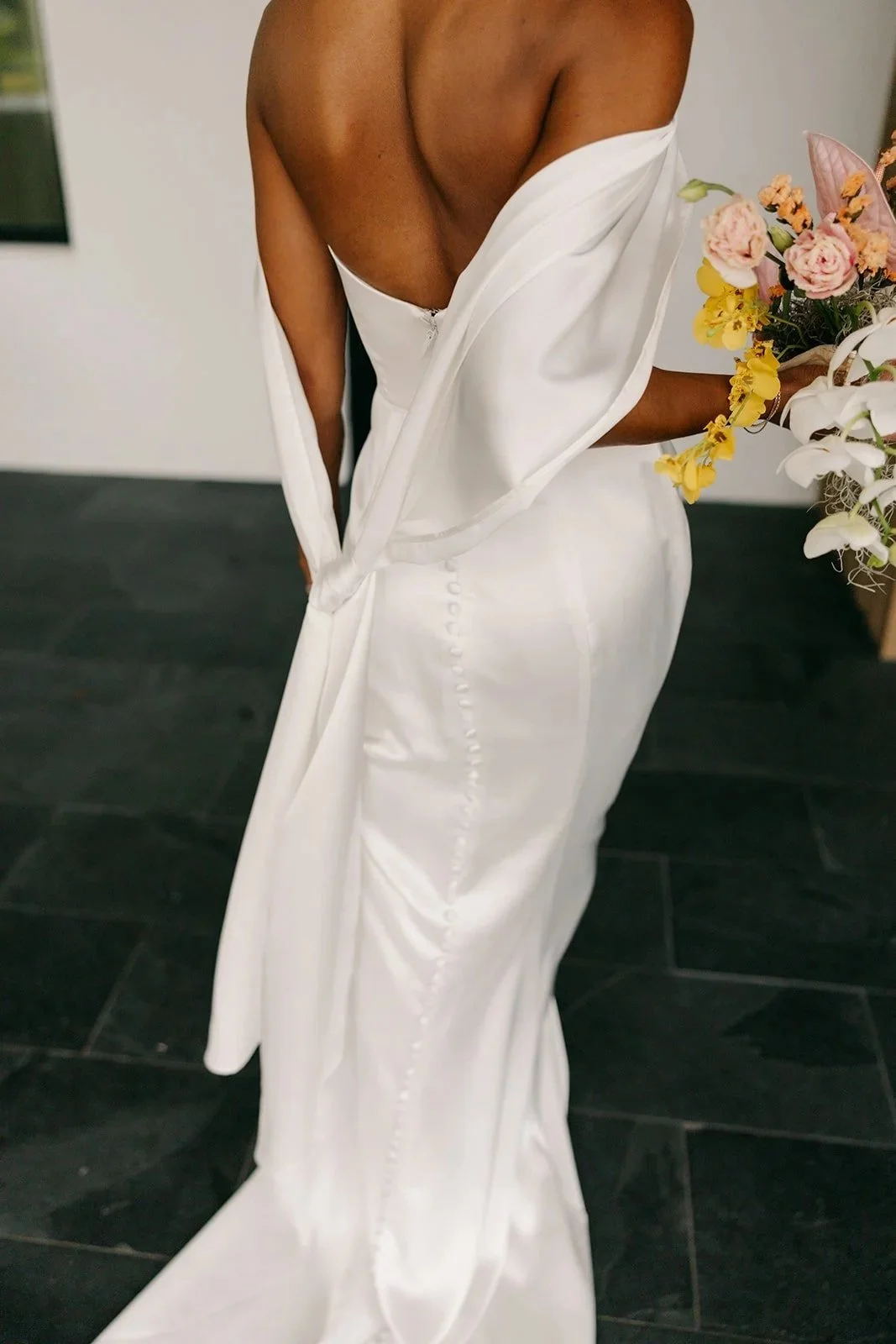 A person in a white dress is holding a bouquet of mixed flowers, with the dress partially unbuttoned at the back, revealing their shoulders and upper back.