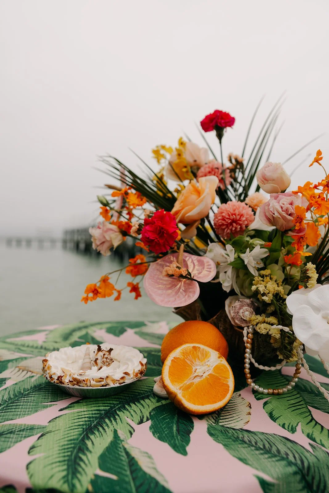 A colorful bouquet of flowers with orange, pink, white, and red blossoms and green leaves, placed on a palm leaf-patterned tablecloth. There are sliced oranges, a full orange, and a dessert topped with whipped cream and almond slices in front of the bouquet. The background appears to be a body of water and a pier.