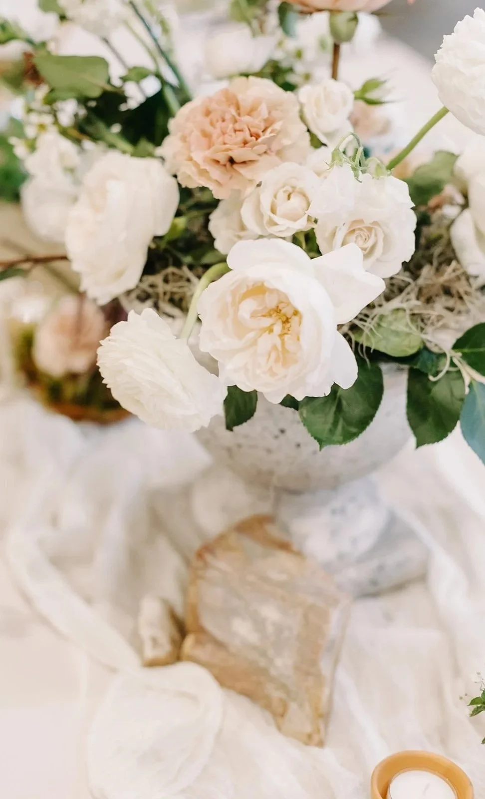 A bouquet of white and pale pink roses in a decorative silver vase, with greenery and moss, placed on a table with a white tablecloth and candle holders.