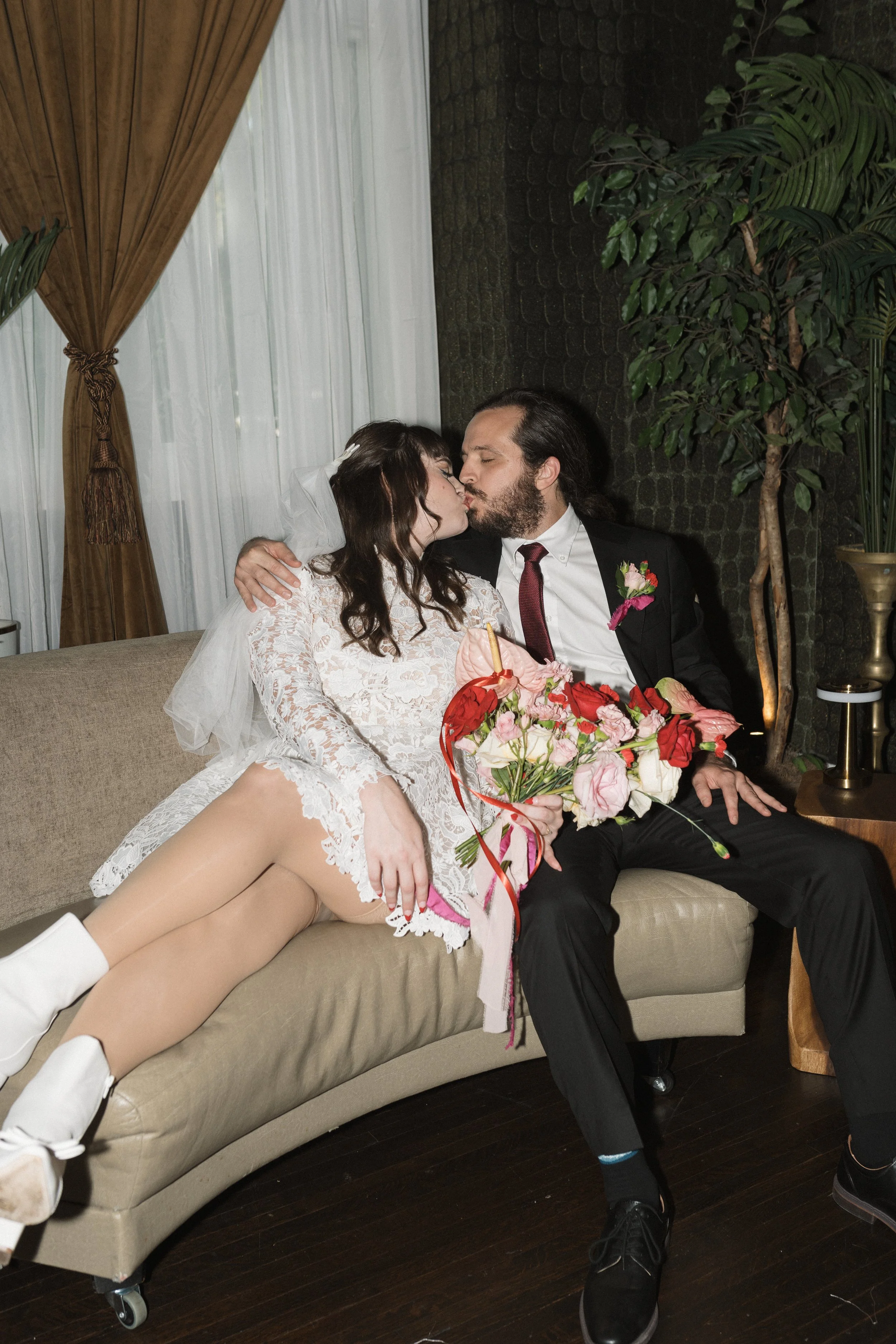 A couple dressed in wedding attire sharing a kiss while sitting on a sofa. The woman is wearing a white lace dress with a veil, and the man is in a black suit with a white shirt and a red tie. They are holding a bouquet of pink and red flowers. The setting appears to be indoors with dark walls, a large potted plant, and drapes with sheer curtains in the background.