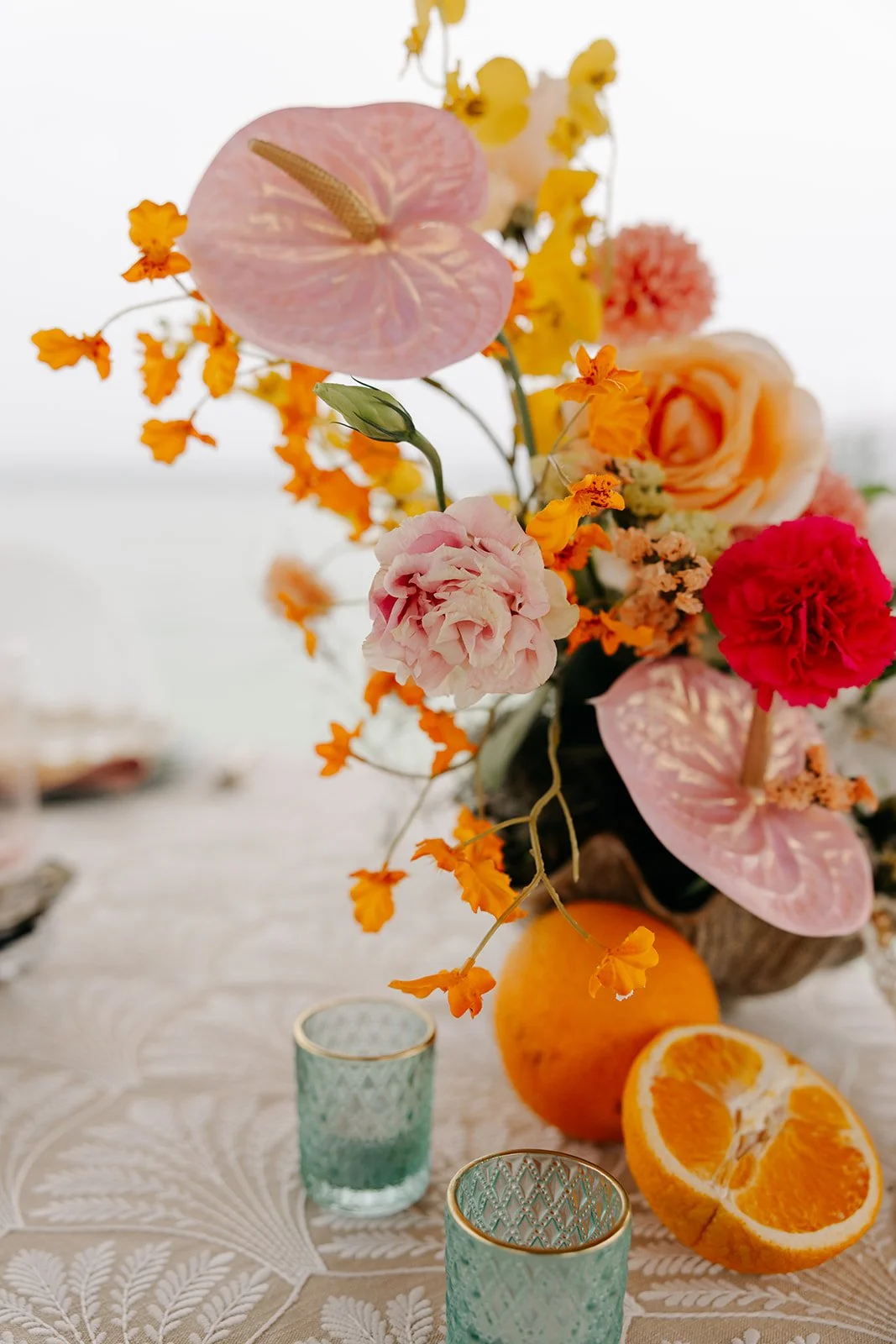 A floral centerpiece featuring pink anthuriums, peach roses, pink carnations, orange marigolds, and other colorful flowers on a table with orange slices, a whole orange, and green candle holders.