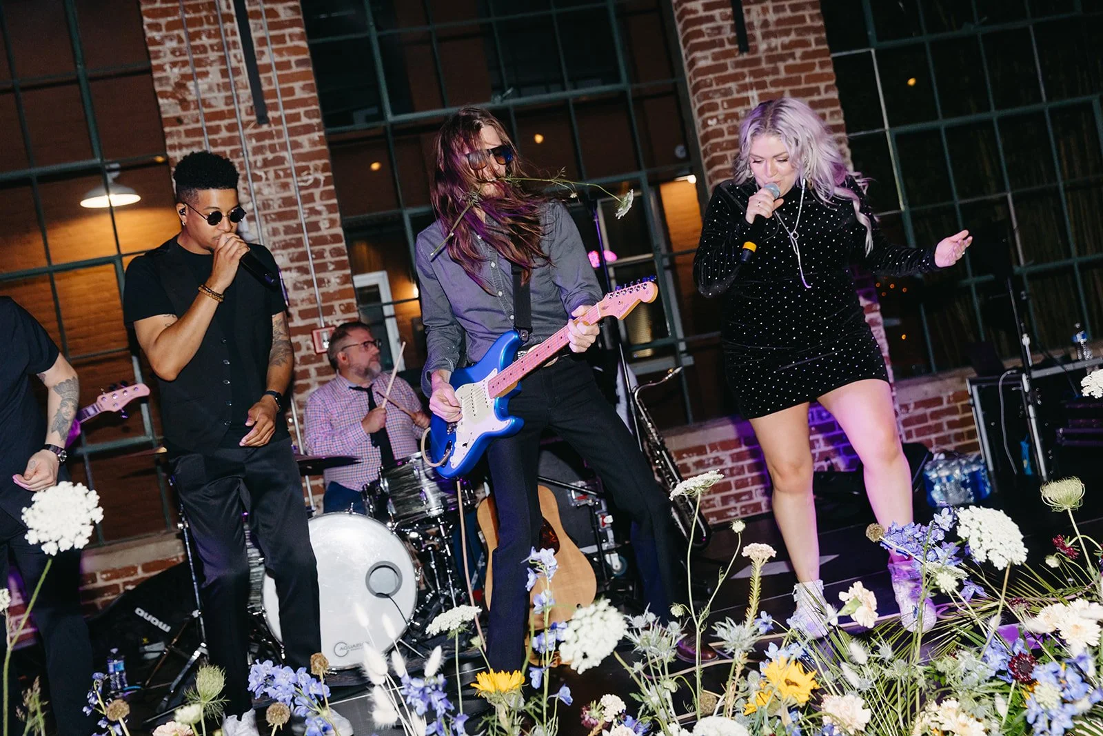 Musicians performing on stage with floral decorations in front, in a venue with brick walls and large windows.