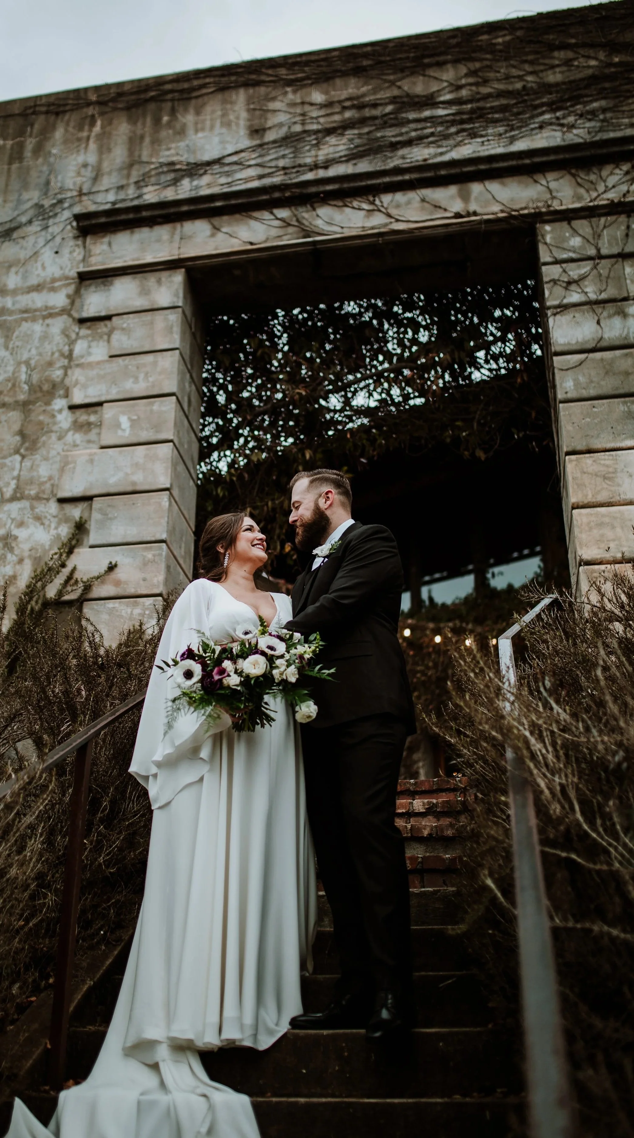 A bride and groom standing on outdoor stairs, gazing at each other, with the bride holding a bouquet of white and purple flowers, in front of a weathered stone building with an archway and climbing vines.