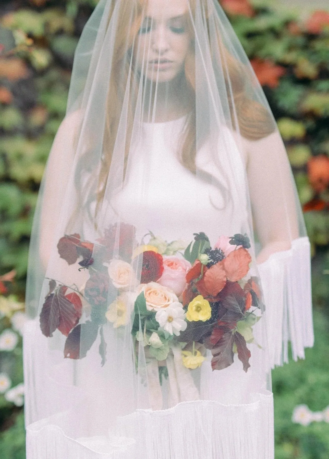 A woman in a white dress is standing outdoors behind a sheer veil, holding a colorful bouquet of flowers, with greenery and blurred flowers in the background.
