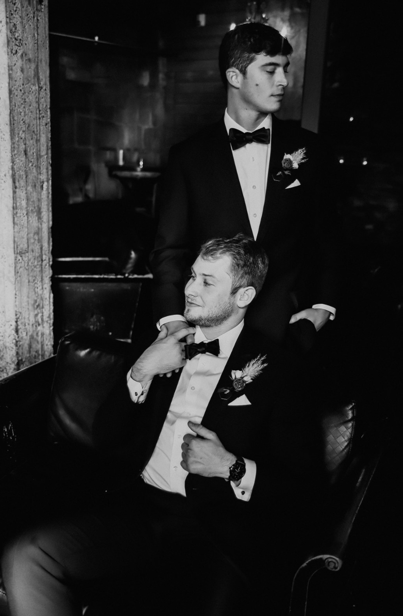 Black and white photo of two men in tuxedos and bow ties, one sitting on a leather couch and the other standing behind him, both with boutonnières on their lapels, in a dimly lit room.