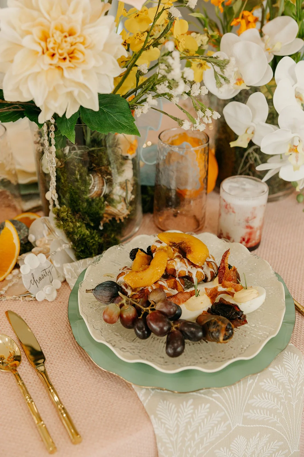 A dessert plate with peaches, grapes, and a half egg, surrounded by decorative flowers and set on a pink tablecloth with gold utensils.