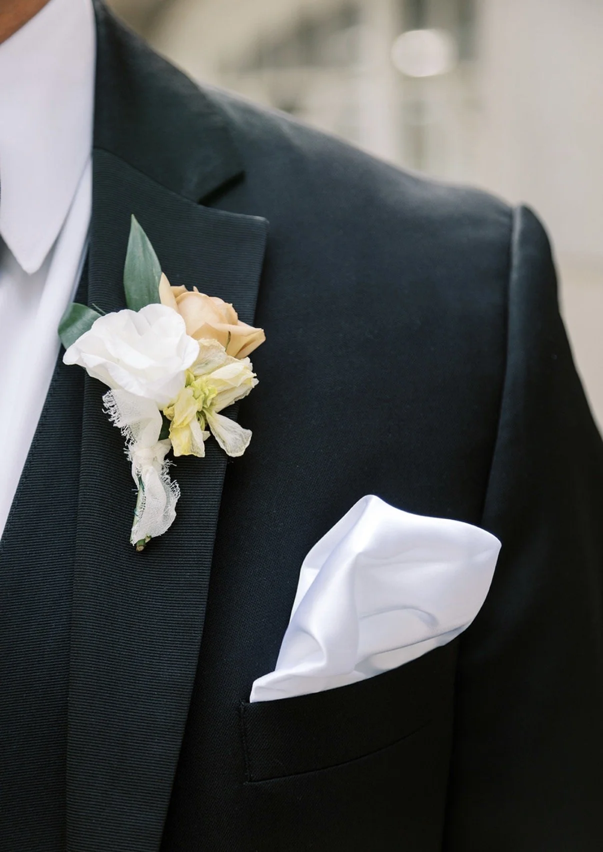 Close-up of a man in a black tuxedo with a white dress shirt, wearing a boutonniere of white and beige flowers with greenery on the lapel and a white pocket square in the breast pocket.