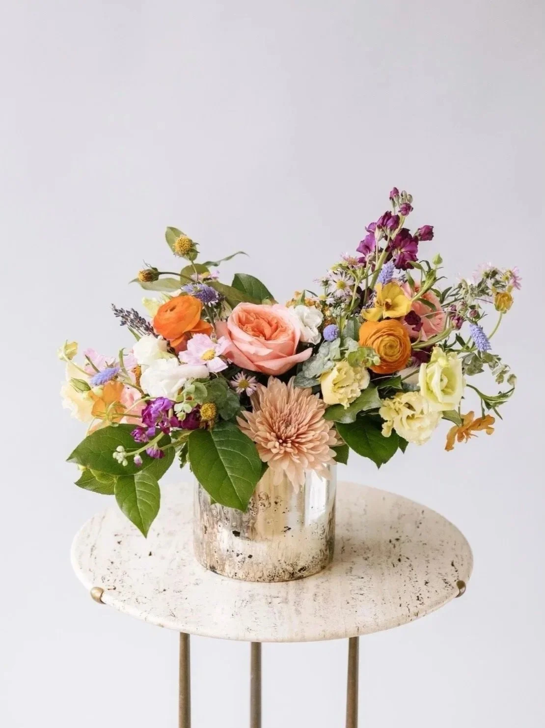 A colorful bouquet of flowers in a reflective silver vase on a small white table.