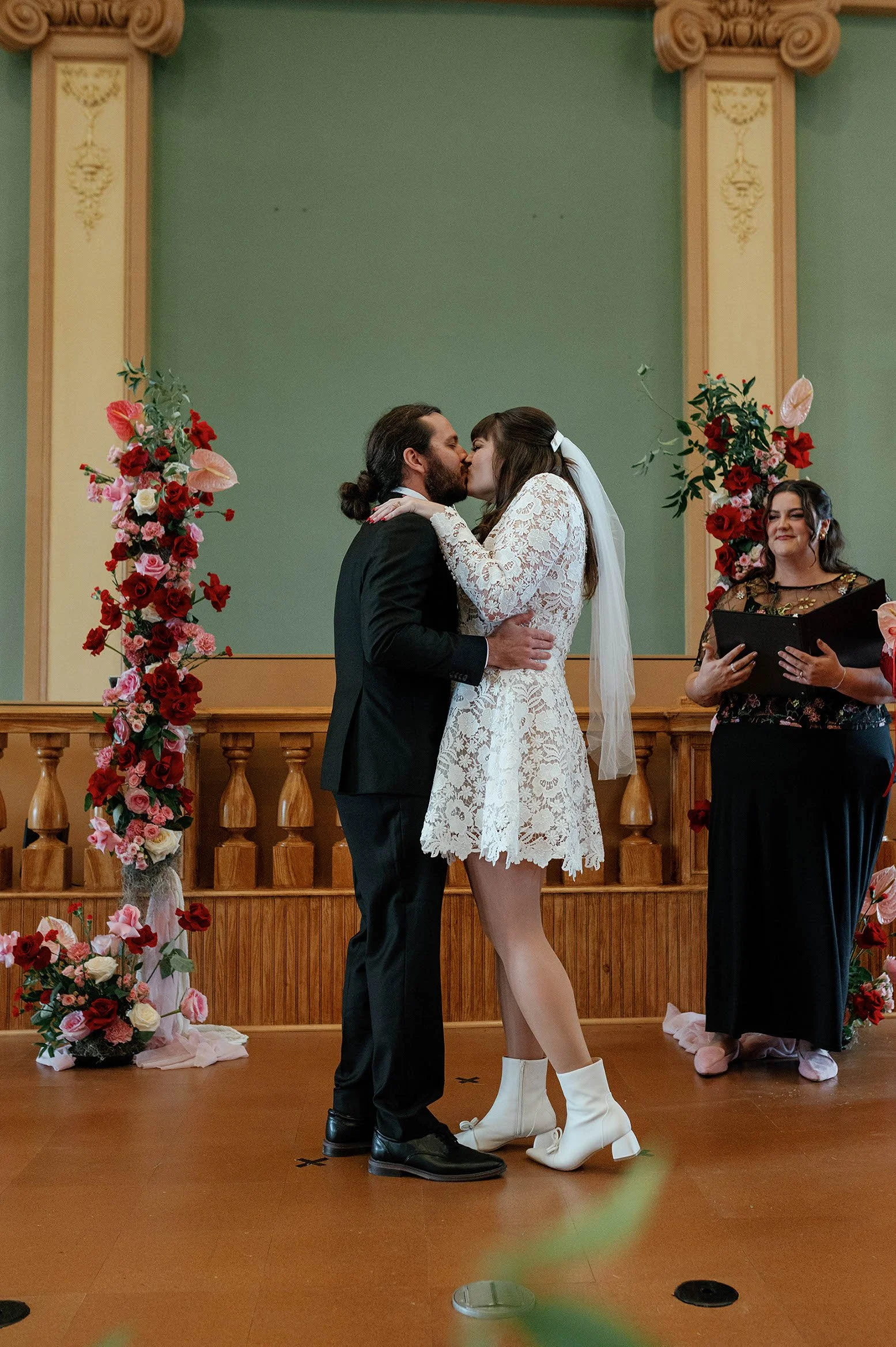 A couple is kissing during a wedding ceremony in front of a decorated floral arch. The bride is wearing a lace dress and white boots, while the groom is in a black suit. An officiant stands to the right, holding a folder.