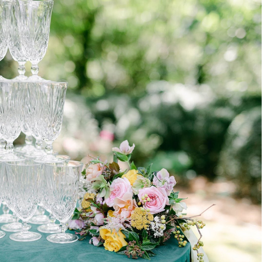 A tiered display of empty crystal champagne glasses next to a bouquet of pink and yellow roses with greenery and small white flowers, outdoors with blurred greenery background.