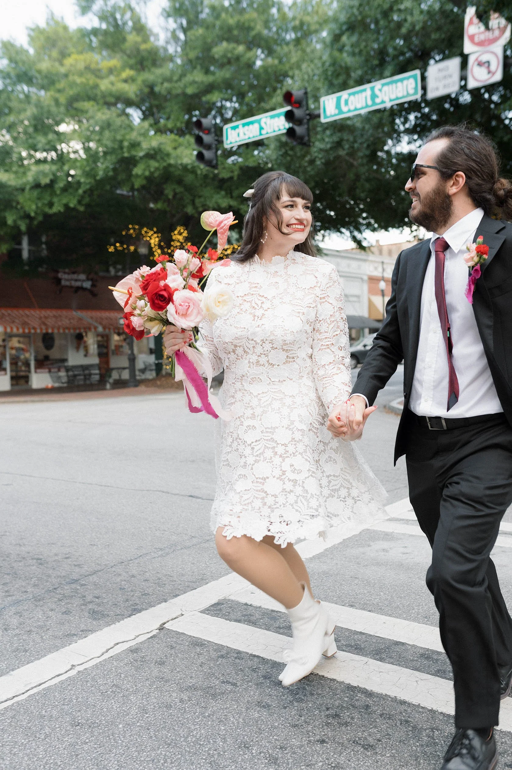 A couple holding hands crossing a street in wedding attire, the woman holding a bouquet and both smiling.
