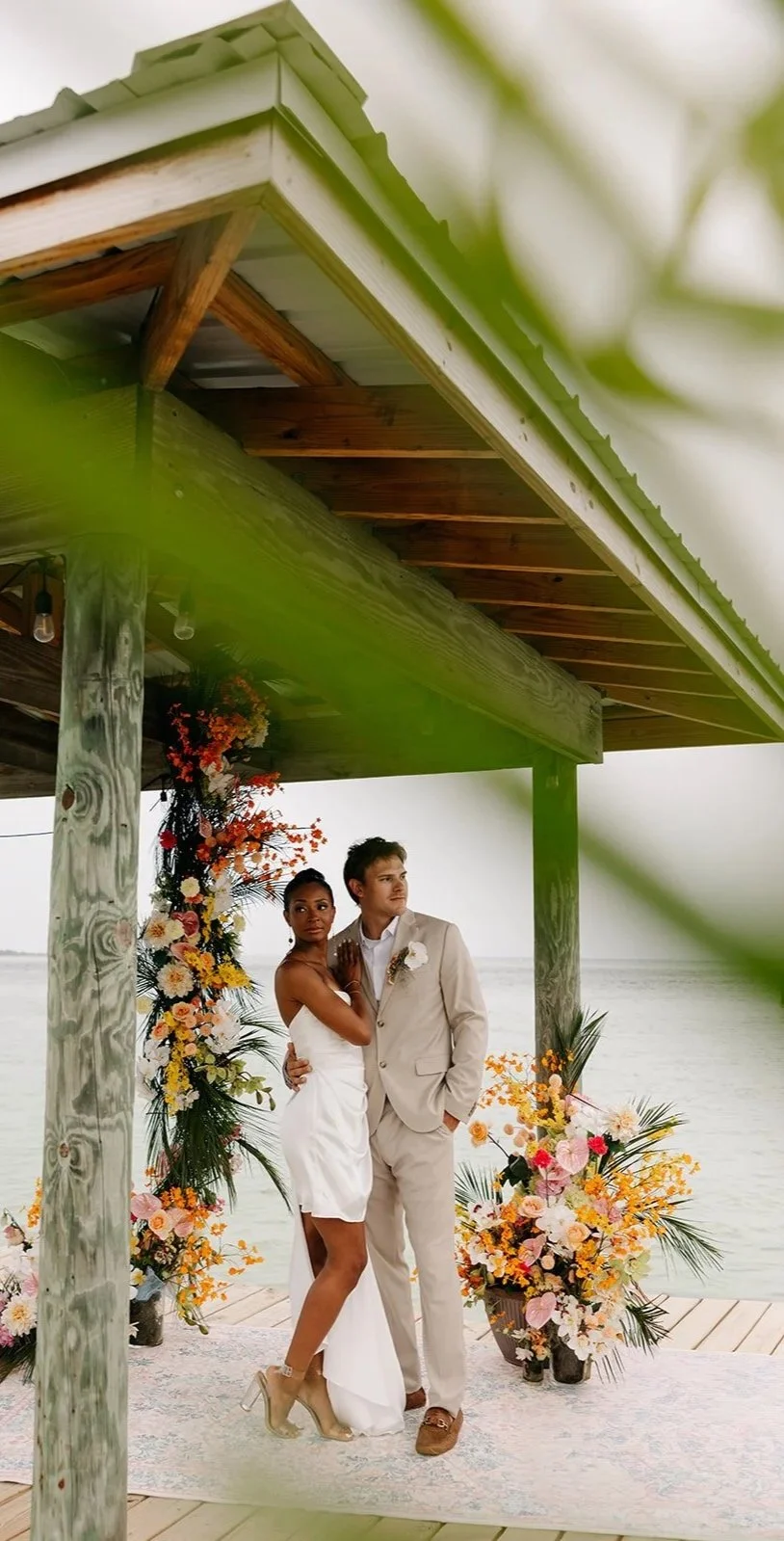 A couple in wedding attire standing under a wooden pavilion with floral decorations by the water.