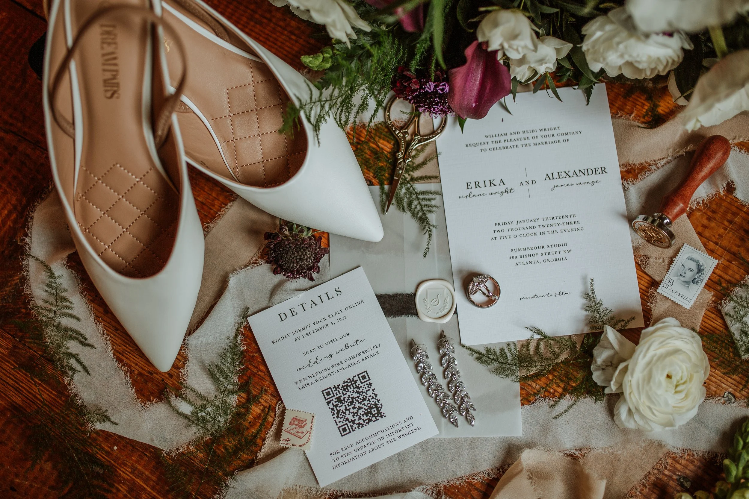 Wedding invitation, white high-heeled shoes, scissors, jewelry, and flowers on a wooden surface.