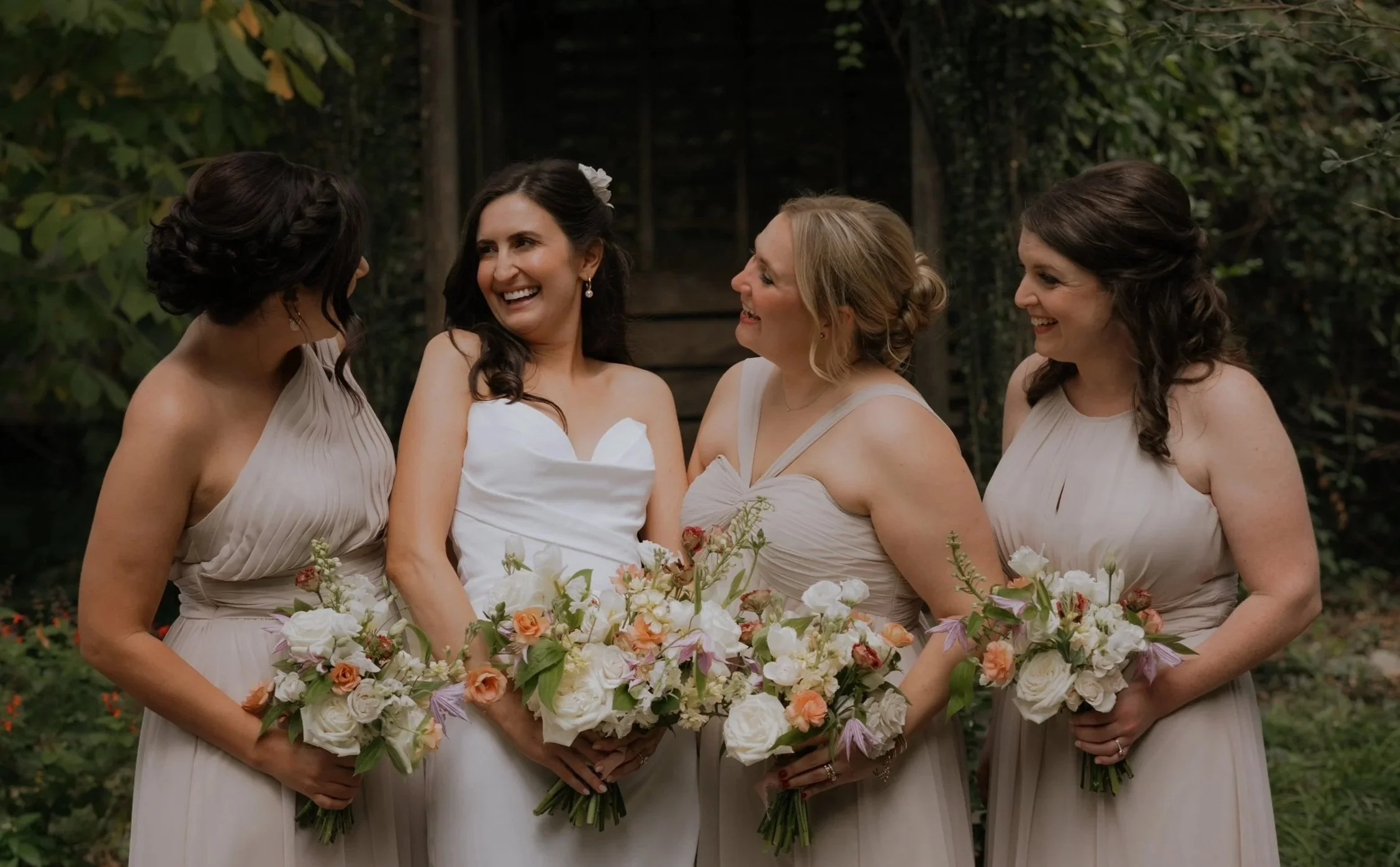 Four women in bridesmaid dresses are gathered outdoors, smiling and holding large bouquets of flowers. One woman in the middle seems to be the bride, wearing a white dress, while the three women around her are bridesmaids in matching beige dresses. They are surrounded by greenery.