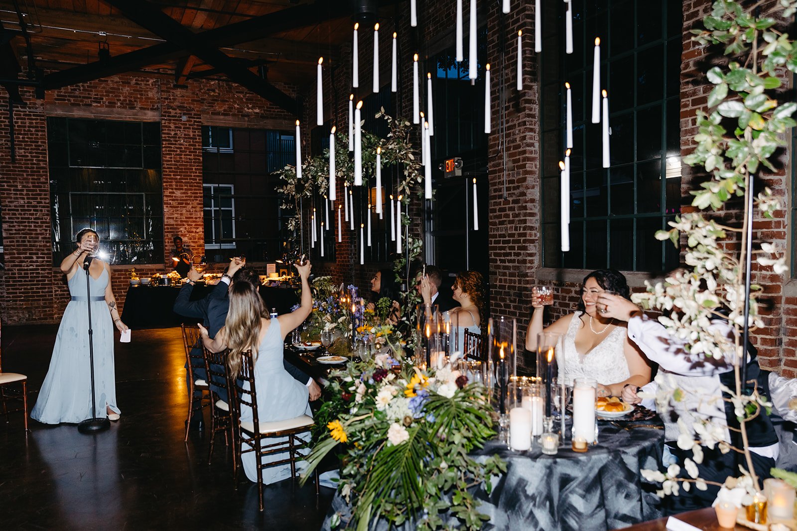 People celebrating at a wedding reception with a seated bride and groom raising glasses, a woman giving a toast, decorative lighting, floral centerpieces, and an industrial-style brick venue.