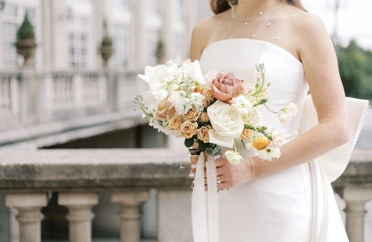 A bride is holding a bouquet of white, blush, and peach-colored flowers while standing on a balcony.