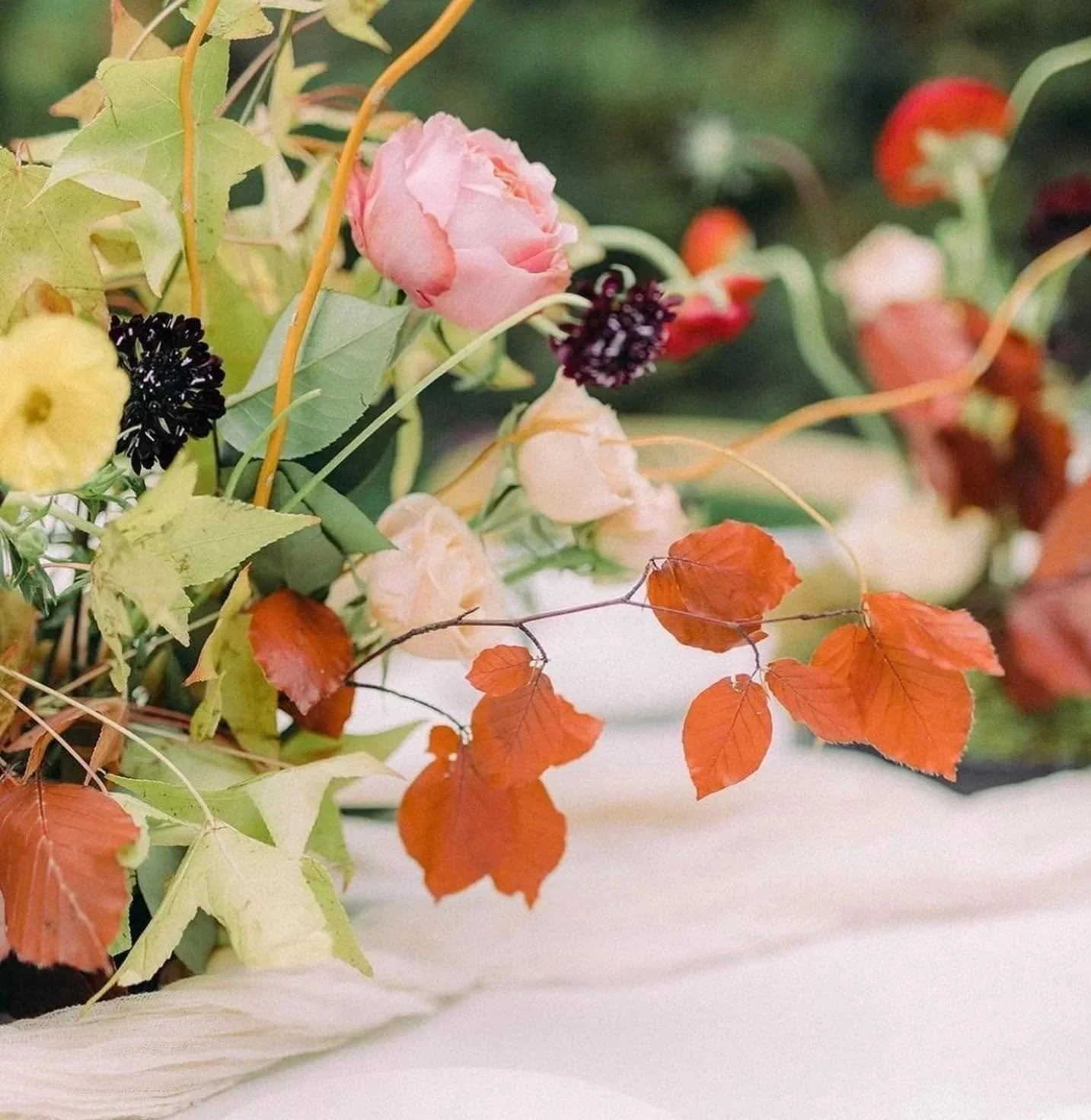 A floral arrangement with pink roses, purple and yellow flowers, green leaves, and orange autumn leaves.
