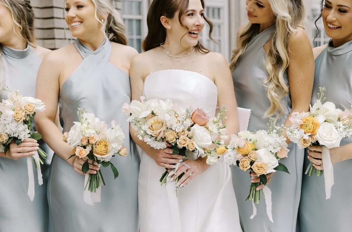 Bride in white wedding dress holding bouquet standing among bridesmaids dressed in light blue dresses, all holding similar bouquets, outdoors in a city setting.