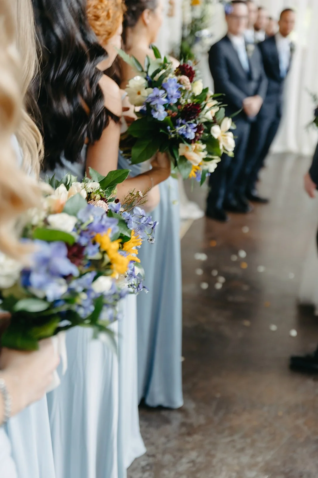 Bridal party holding bouquets of flowers during wedding ceremony with line of groomsmen in suits in the background.