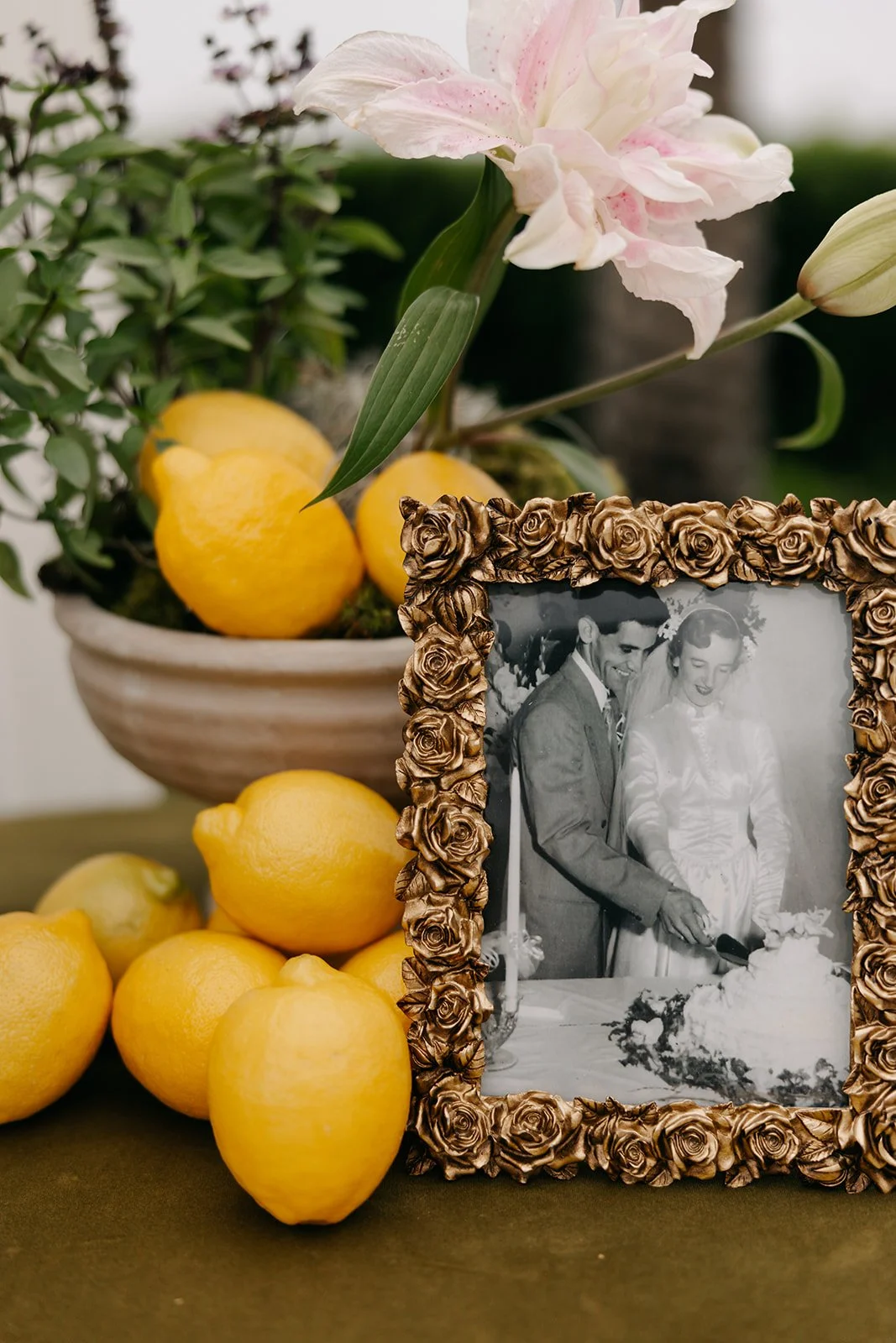 Yellow lemons in a ceramic bowl, a framed black-and-white wedding photo with a rose pattern frame, pink lilies, green leaves, and stems.