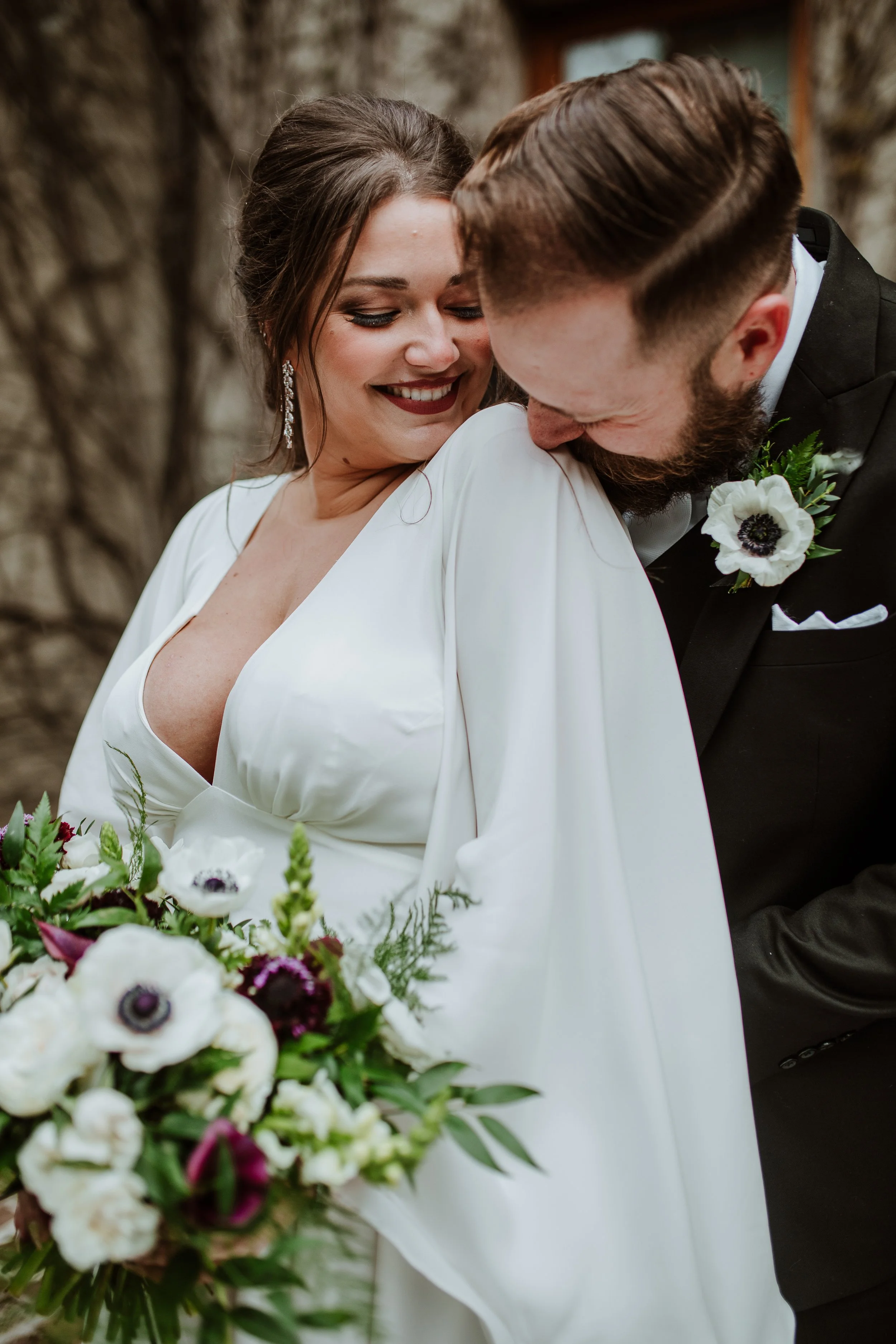 A bride and groom share a close, joyful moment during their wedding, with the bride smiling and the groom gently resting his forehead on her shoulder. The bride wears a white wedding dress and earrings, while the groom is in a black tuxedo with a boutonnière. There is a bouquet of white and dark purple flowers in the foreground.