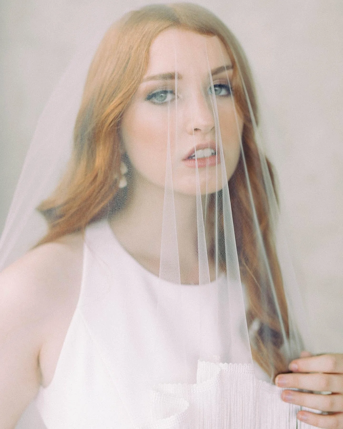 A woman with red hair and blue eyes wearing a white dress and a bridal veil, holding a smartphone in her left hand.