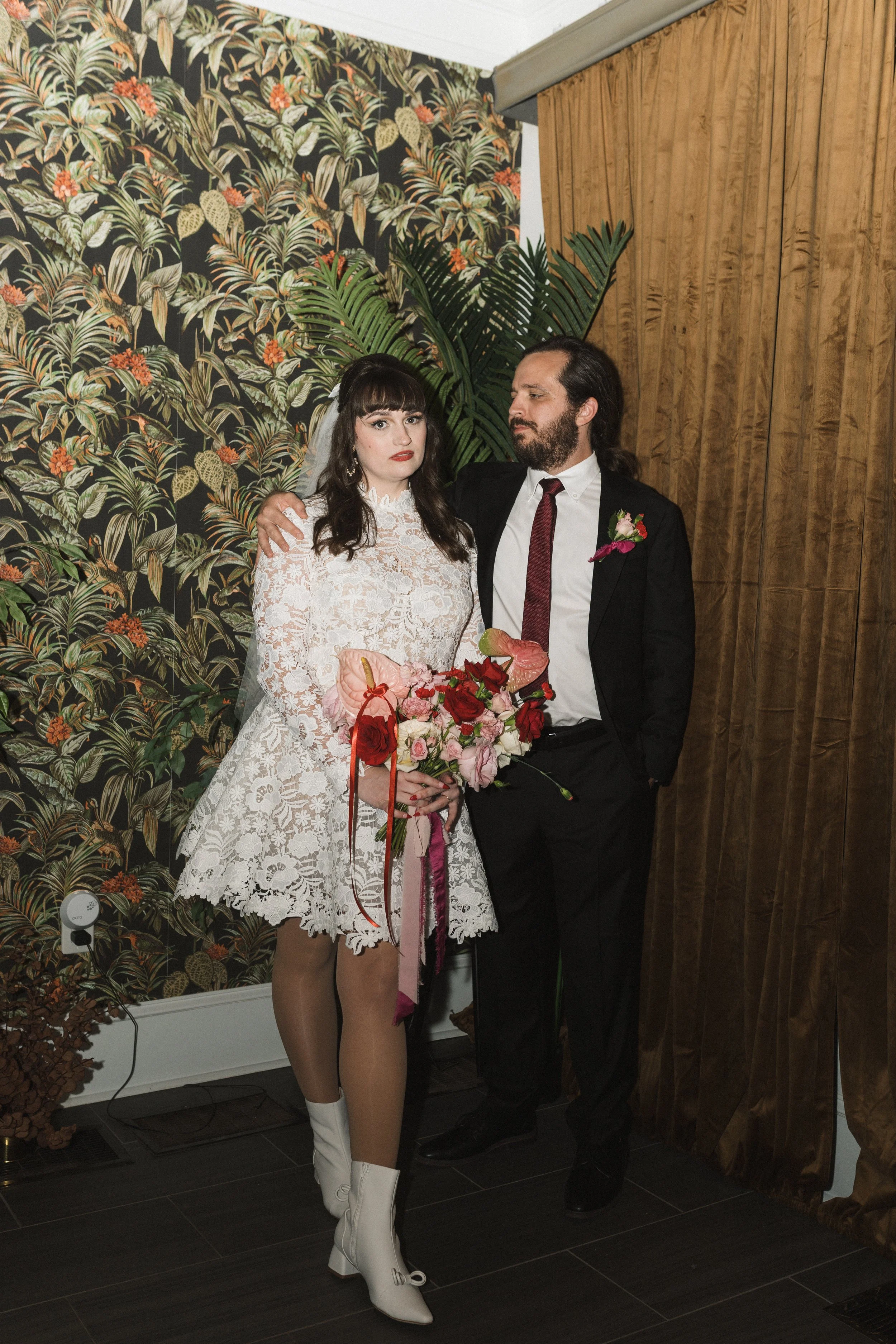 A couple standing indoors, dressed in wedding attire, with the woman wearing a lace wedding dress and white boots holding a bouquet of flowers, and the man in a black suit with a white shirt and dark red tie, against a backdrop of tropical patterned wallpaper and a wooden wall.