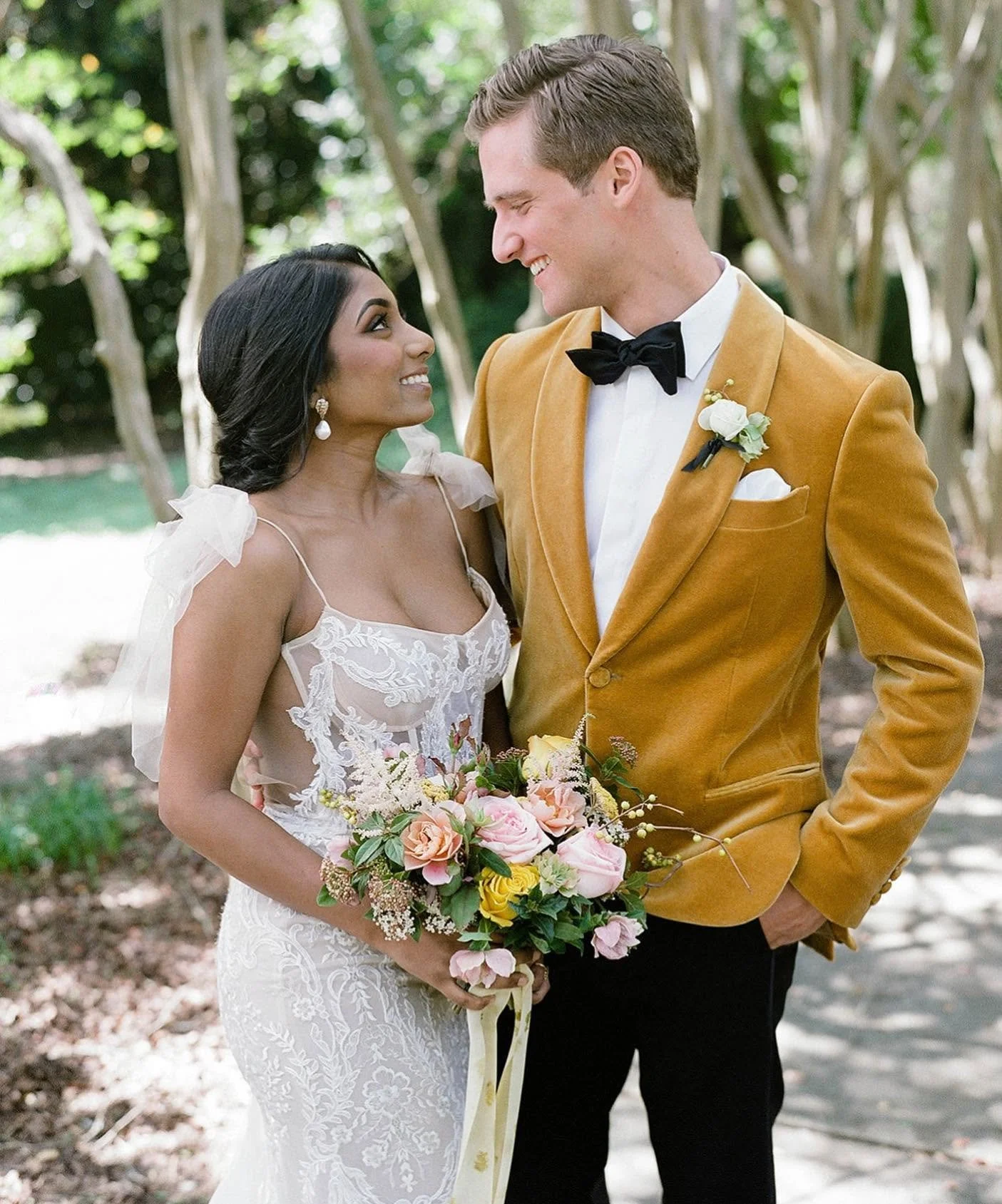 A newlywed couple standing outdoors on a sunny day, gazing into each other's eyes, with trees in the background. The bride is holding a bouquet of pink, yellow, and white flowers, and wearing a lace wedding dress with tulle shoulder details. The groom is dressed in a mustard-colored suit jacket, white shirt, black bow tie, and has a white boutonniere on his left lapel.
