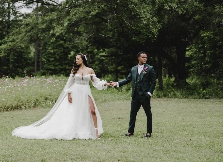 A bride and groom standing on a grassy field, holding hands, with trees in the background. The bride wears a white wedding gown with off-shoulder sleeves and a thigh-high slit, and the groom wears a dark suit with a tie and boutonniere.