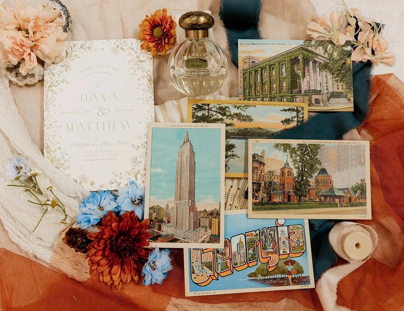 A wedding invitation surrounded by artificial flowers, vintage postcards of New York City landmarks, a bottle of perfume, and decorative fabrics in peach, cream, and teal colors.