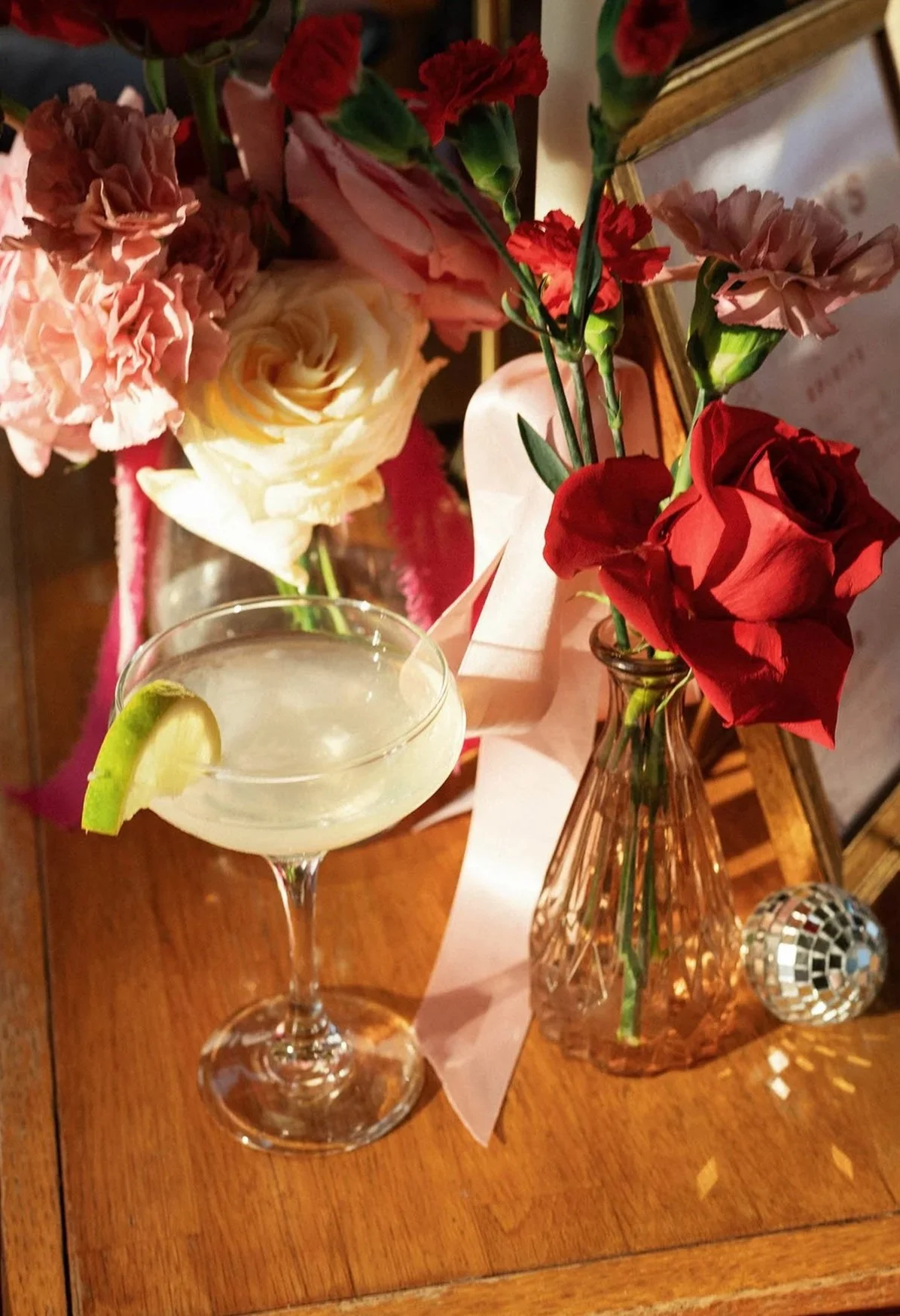 A cocktail with a lime wedge in a glass, positioned next to a bouquet of pink, cream, and red flowers in a glass vase, with a small disco ball on a wooden table.