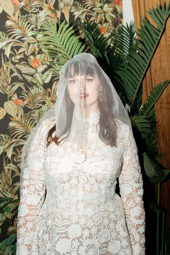 A woman dressed in a white lace dress with a veil covering her face, standing in front of tropical wallpaper and green plants.