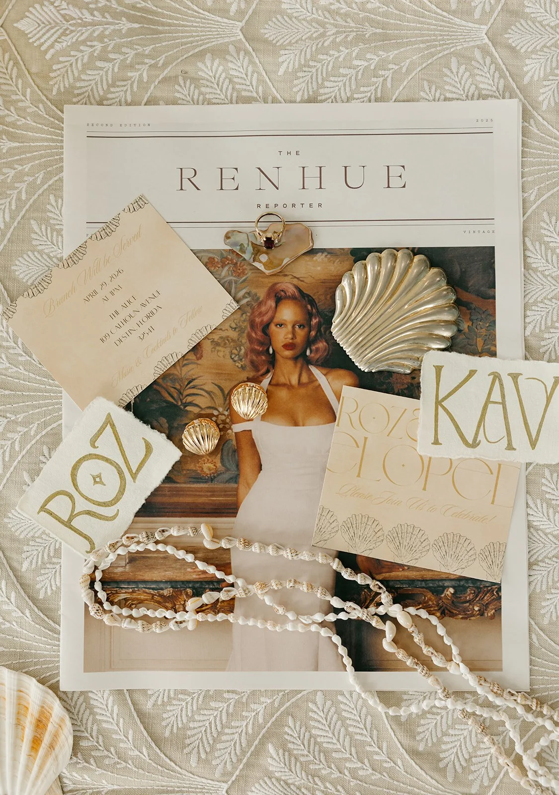 A collection of jewelry and paper items on top of a magazine page featuring a woman in a white dress, with decorative shell accessories, a pearl necklace, and cards with gold lettering spelling 'ROX', 'KAY', and other text, all placed on textured white fabric.