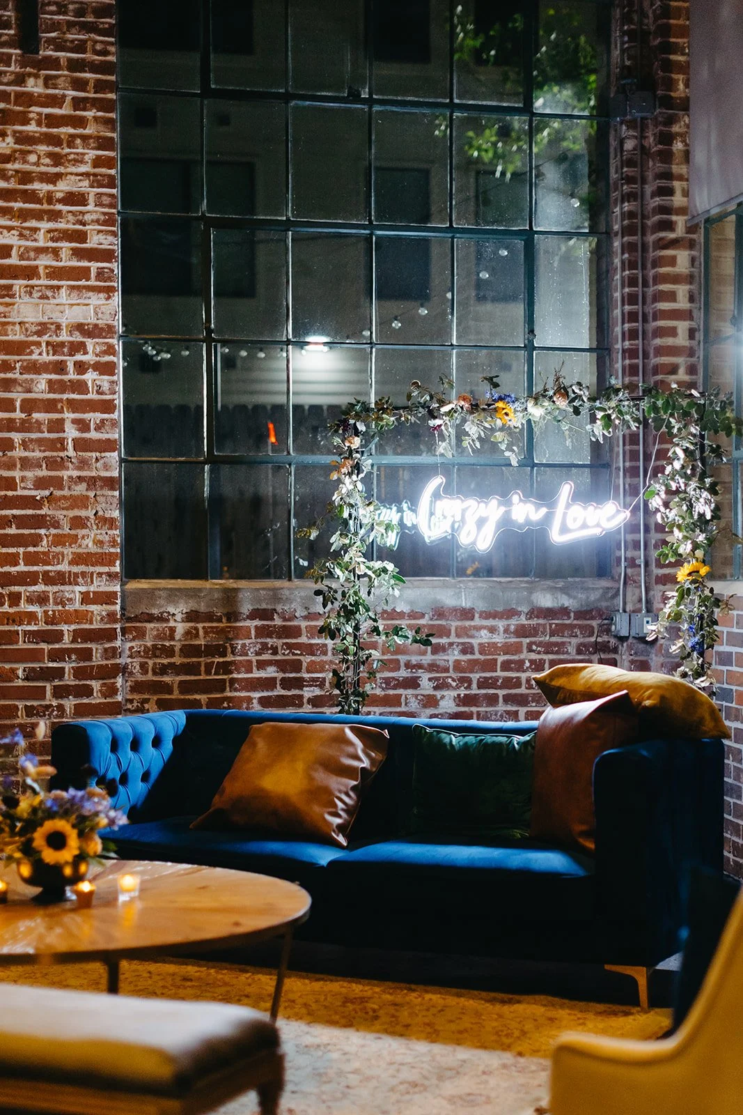 A cozy indoor space with a brick wall and large window. There is a neon sign that says 'Love in Love', a dark blue sofa with yellow and dark green pillows, a wooden coffee table with a sunflower centerpiece, and decorative flowers on a frame around the neon sign.