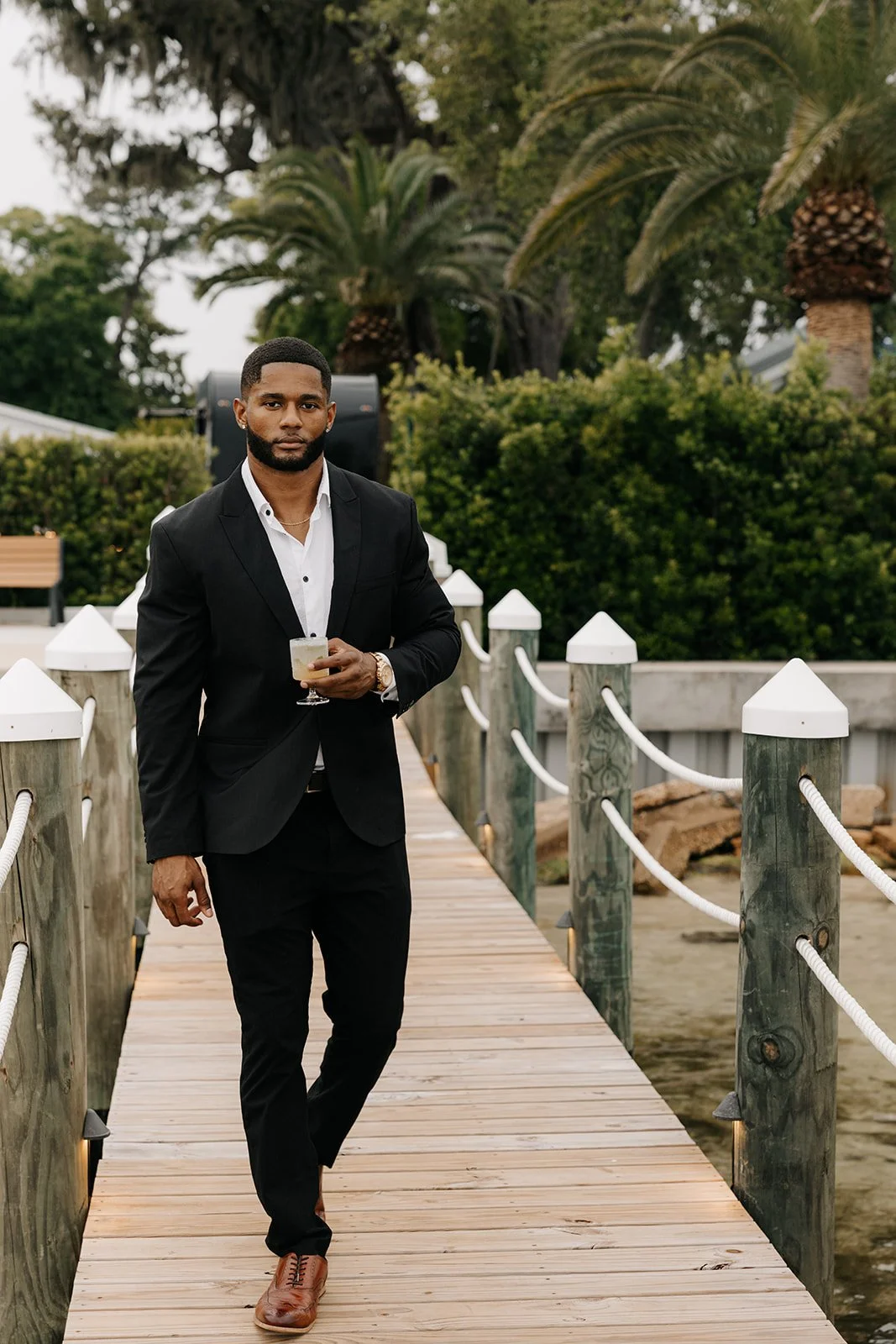 A man in a black suit and white shirt holding a cocktail, walking along a wooden dock with white posts and ropes, surrounded by green palm trees and bushes.