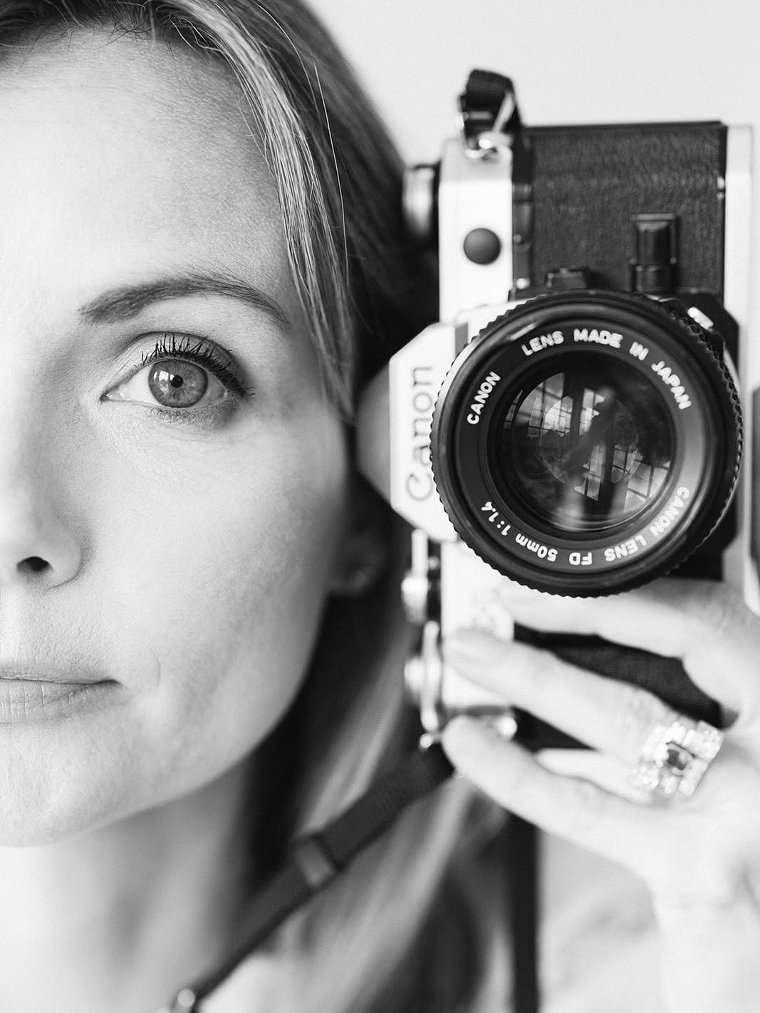 A close-up black and white photograph of a woman with light skin and bright eyes, holding a Canon camera with the lens pointed toward a mirror, capturing her reflection.