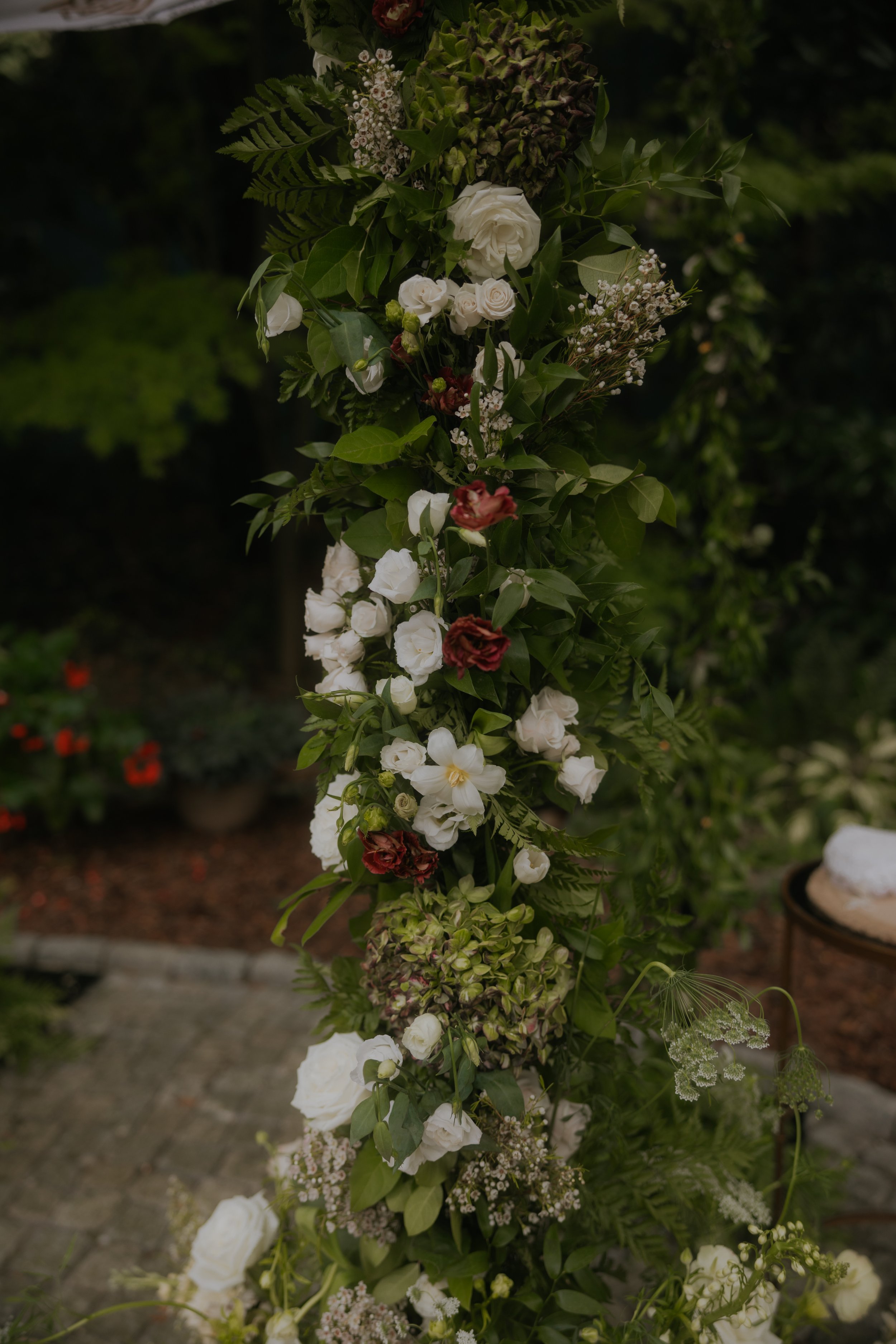 A vertical floral arrangement with white roses, green ferns, and small white flowers, set outdoors with a blurred background of greenery and a brick walkway.