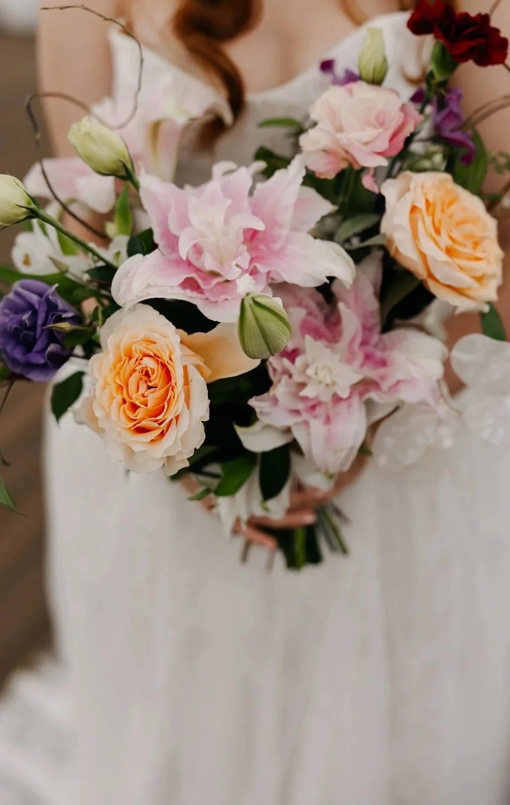 A close-up of a colorful bouquet of flowers featuring roses, lilies, and other blooms in shades of peach, pink, purple, and white.