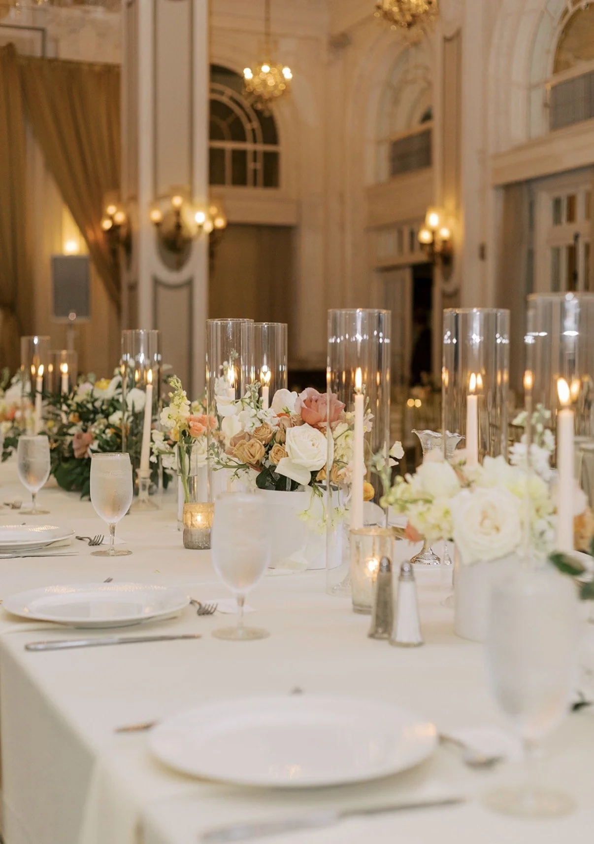 Elegant banquet table with white tablecloth, floral centerpieces, and tall candles in glass holders, set in a grand, ornate room with chandeliers and arched windows.