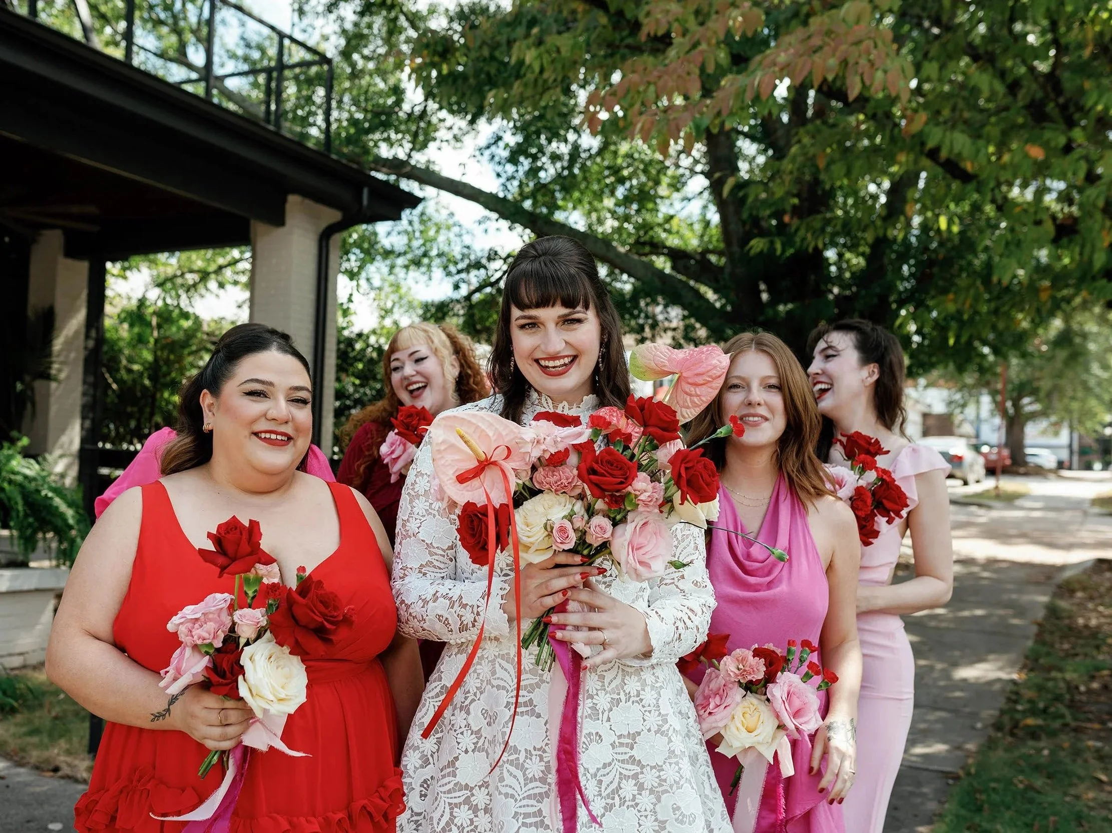 A group of women dressed in pink and red dresses holding bouquets of pink, red, and white flowers, gathered outdoors with trees and a building in the background.