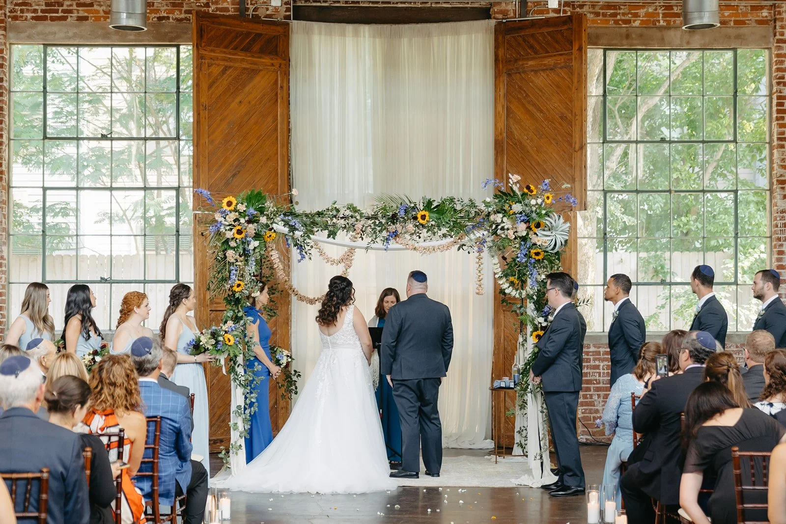 Indoor wedding ceremony with a bride and groom standing before officiant, decorated with floral arch, surrounded by bridesmaids and groomsmen, guests seated, large windows, brick walls, wooden backdrop, and greenery outside.