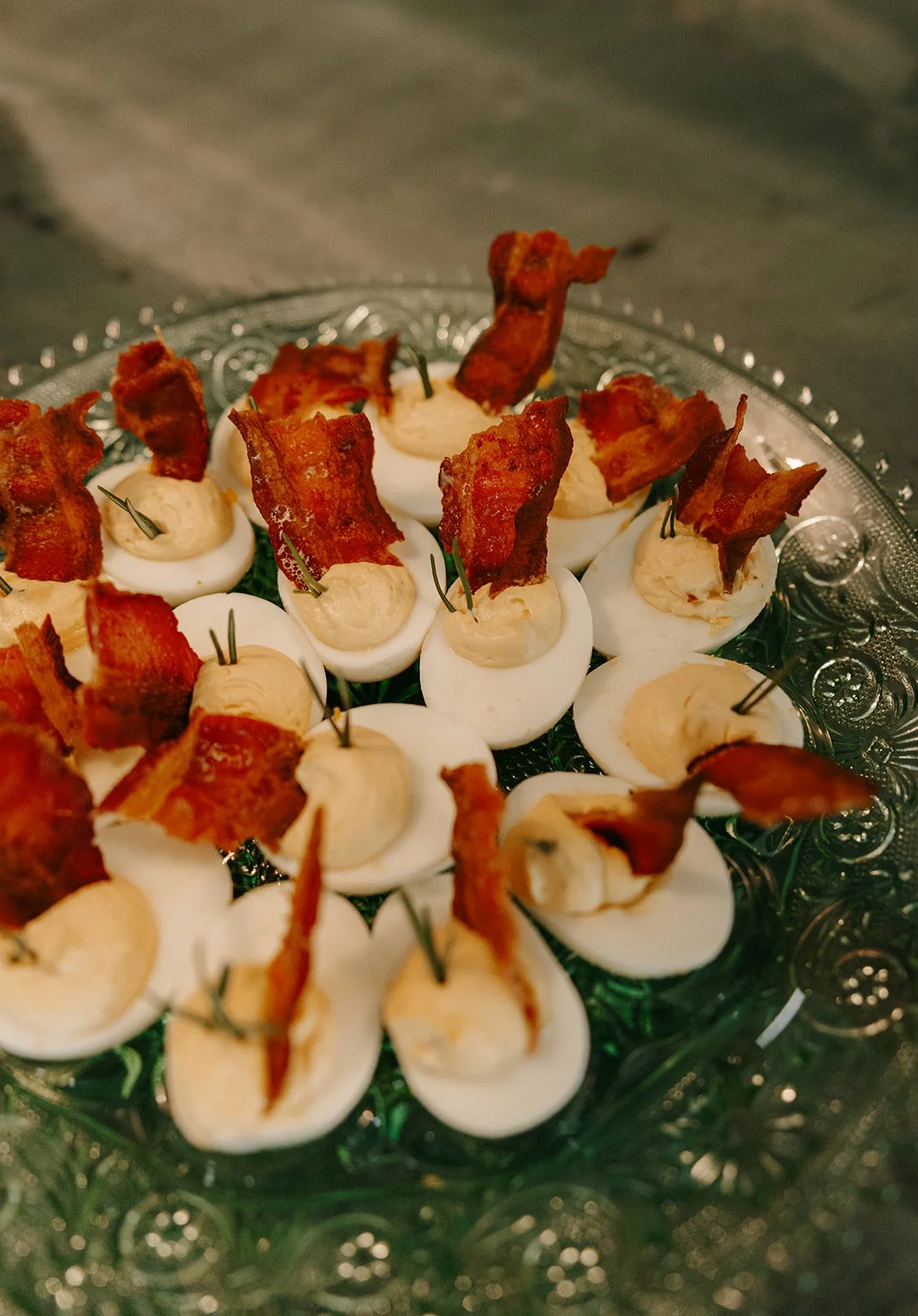 Deviled eggs topped with bacon and small garnishes on a decorative clear glass tray