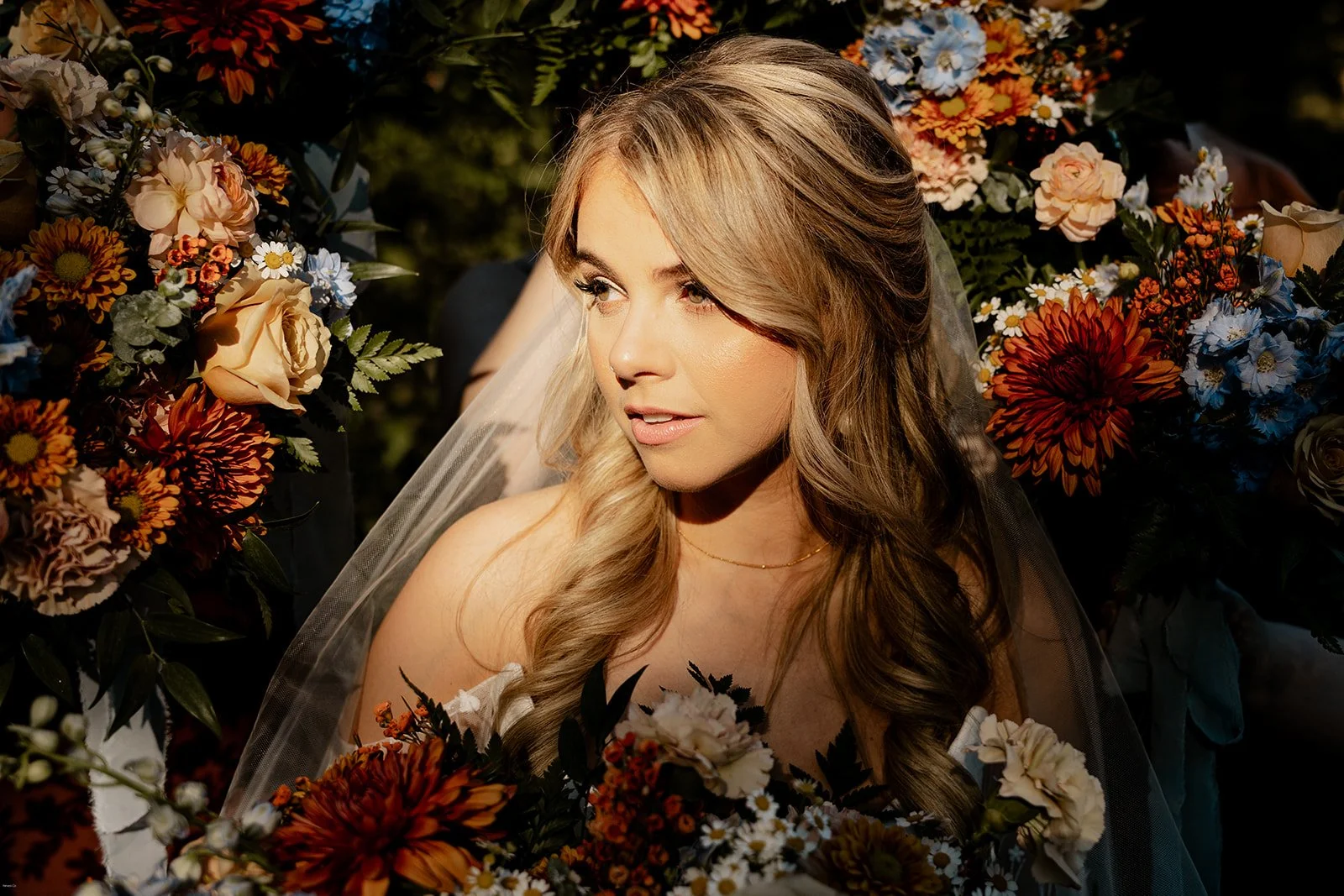 A woman with long, blond wavy hair, wearing a wedding veil, is surrounded by colorful flowers.