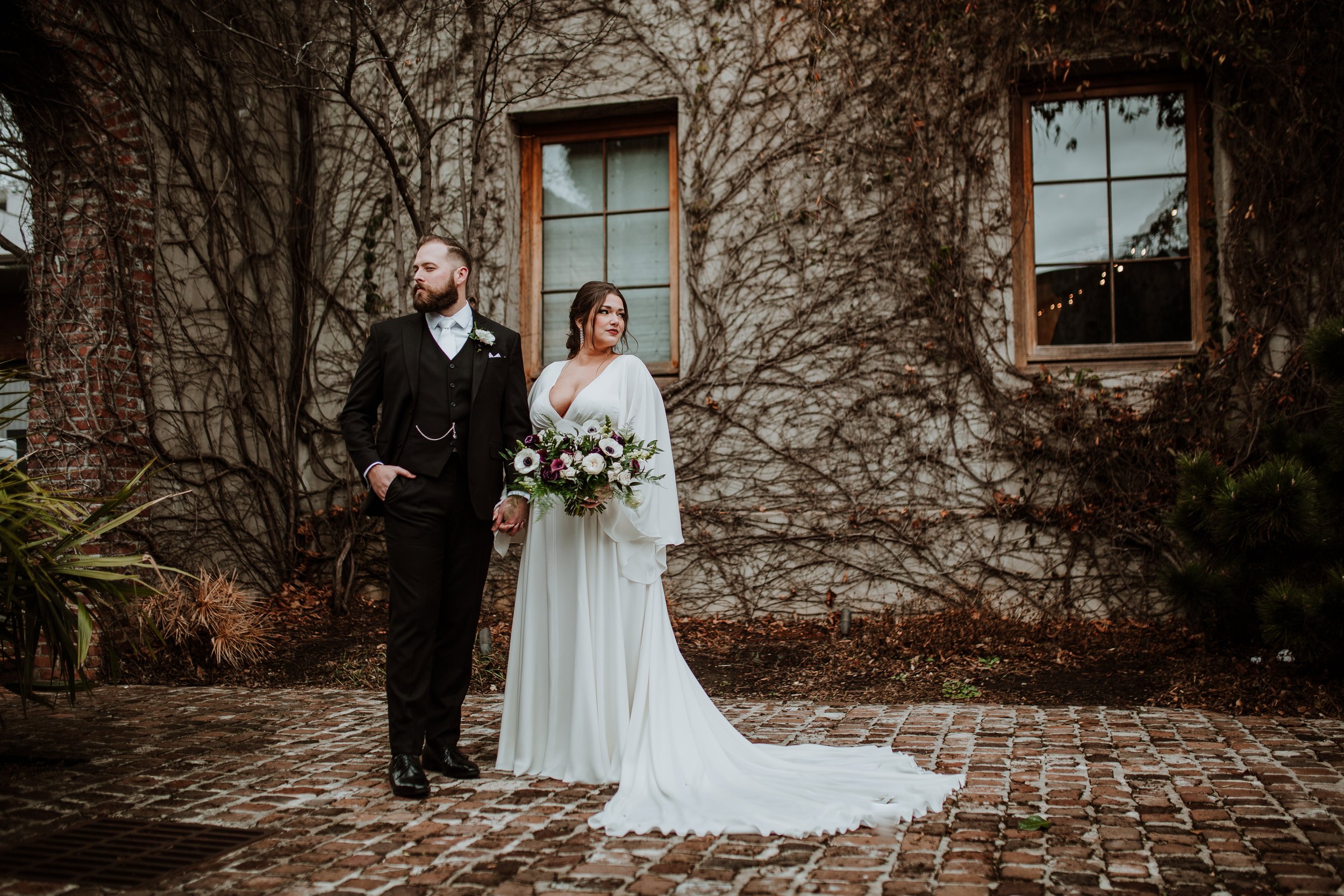 A bride and groom standing outdoors in front of a rustic wall with bare vine branches. The bride holds a bouquet and wears a white dress, while the groom is dressed in a black tuxedo with a white shirt and bow tie.