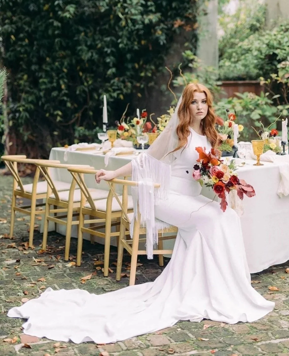 A woman in a white wedding dress and veil is sitting outdoors at a table with a floral arrangement, surrounded by greenery.
