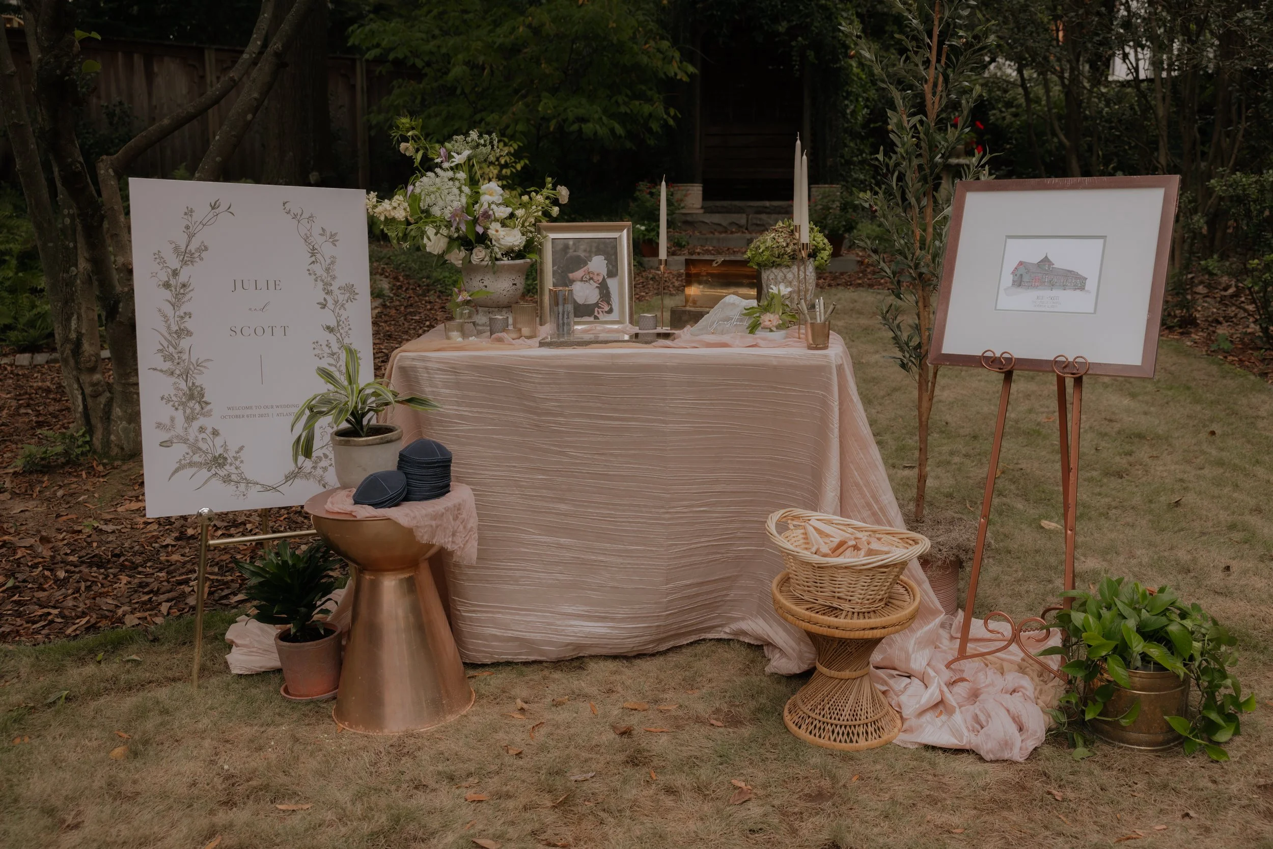 An outdoor wedding reception table decorated with flowers, framed photos, candles, and a sign with the names Julie and Scott, set in a garden with trees and greenery.
