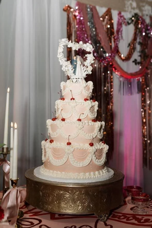 A multi-tiered wedding cake decorated with white icing and cherries, topped with a bride and groom figurine, on a round ornate table at a celebration with pink and gold decorations in the background.
